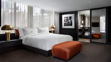 A modern hotel room with a large bed, a sitting area with a red ottoman, and a large window offering a city view.