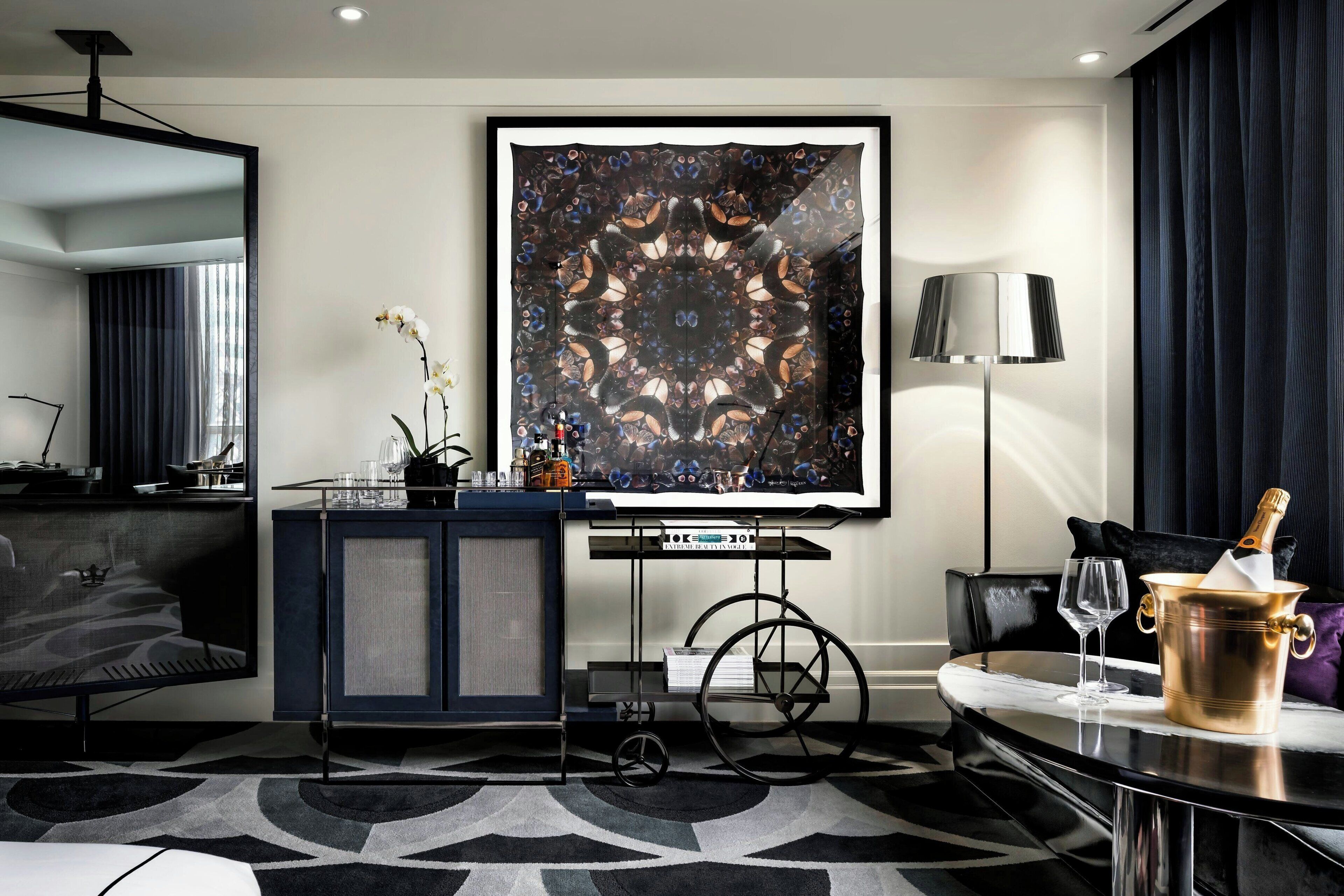 Una habitación de hotel moderna con una gran obra de arte abstracta, un minibar, un escritorio con una computadora portátil y una zona de comedor con una mesa preparada para dos.