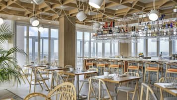 A modern restaurant with high ceilings, exposed beams, and large windows offering city views. Tables are arranged with wicker chairs, and a bar area is visible with bottles and glasses.