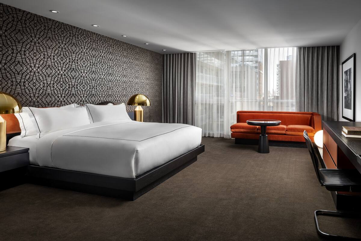 A modern hotel room with a large bed, a sofa, a small table, and a view of the city through large windows.