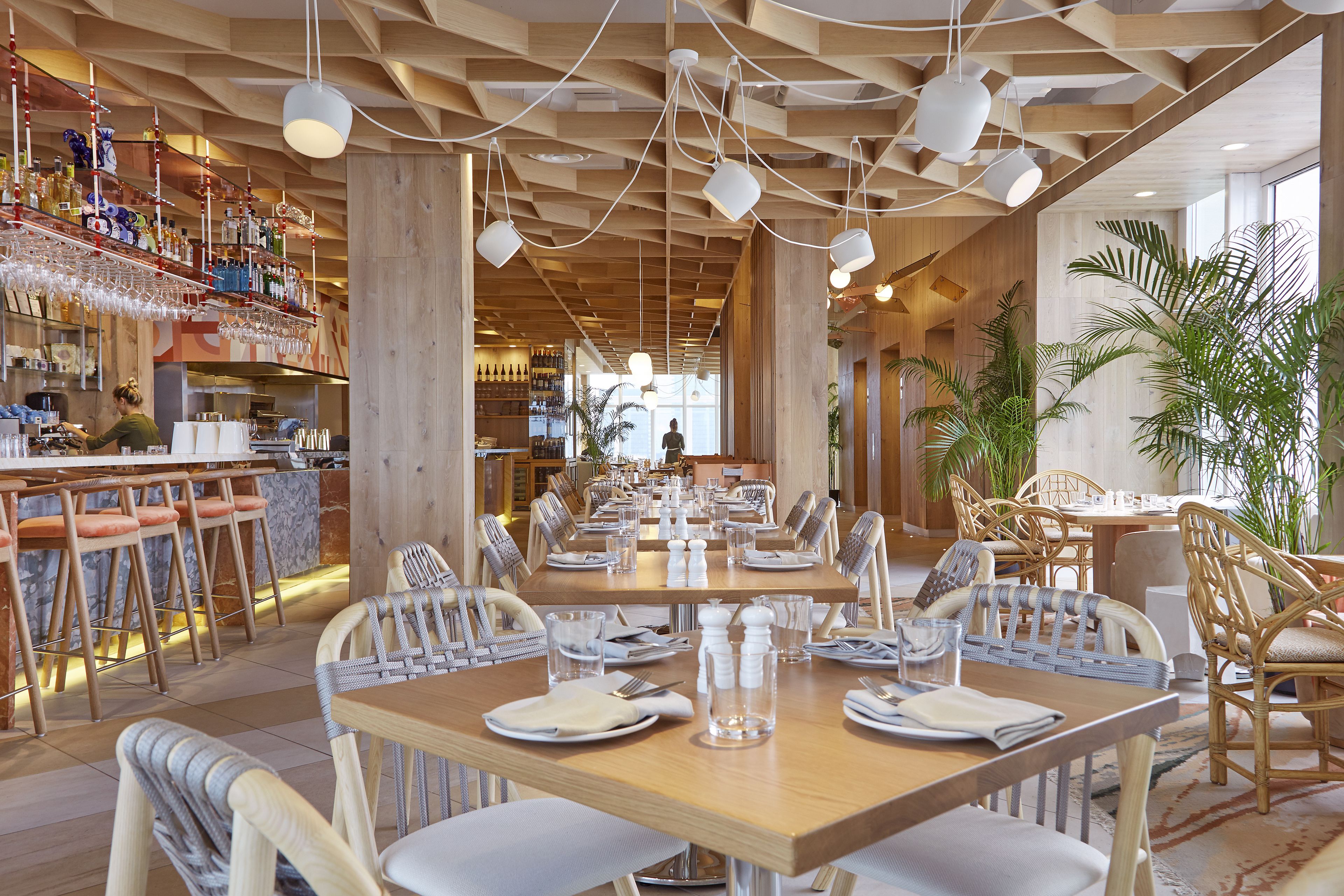A restaurant with wooden tables and chairs, a bar area with shelves of bottles, and a view of the city through large windows.