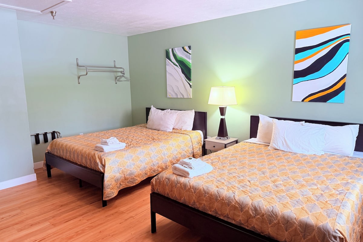A hotel room with two beds, a nightstand with a lamp, a wall art piece, and a ceiling light fixture.
