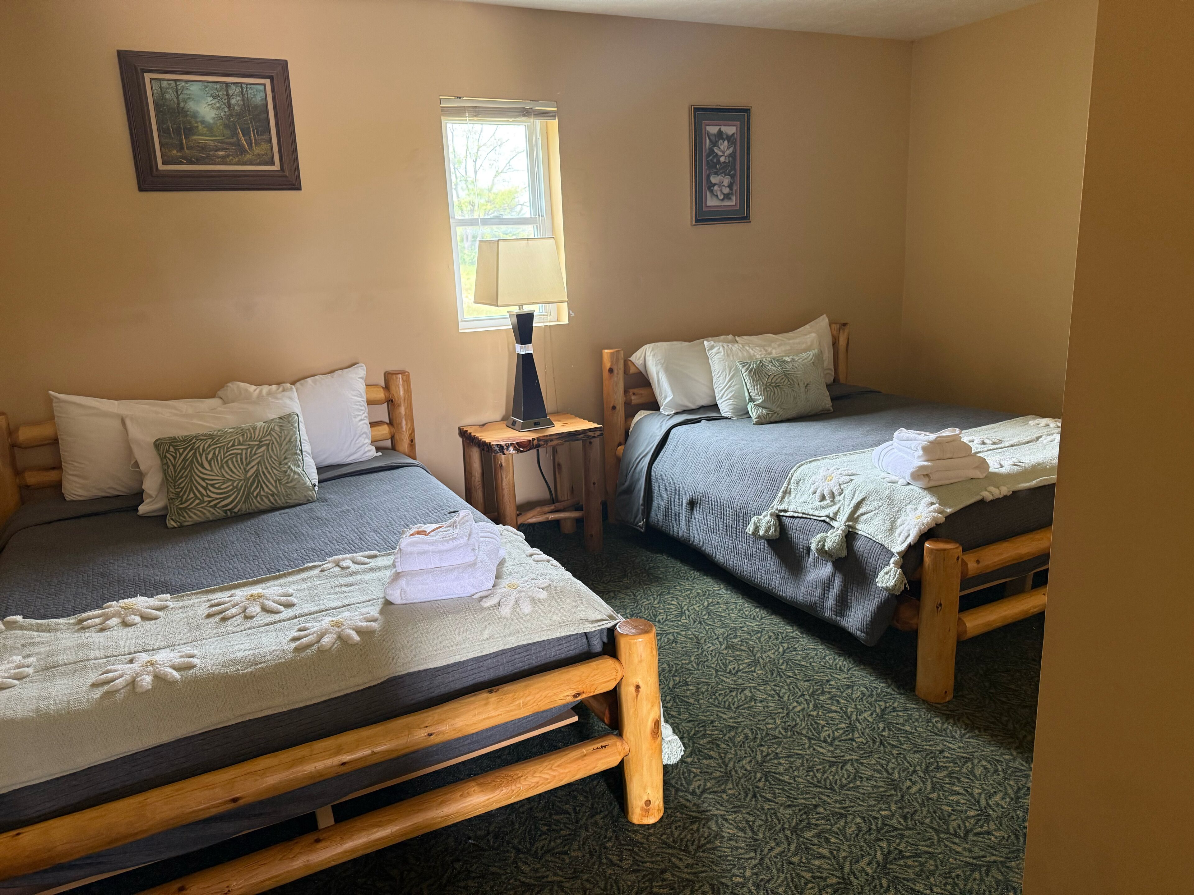 Two wooden beds with grey bedding, a small table with a lamp, and framed pictures on the walls.