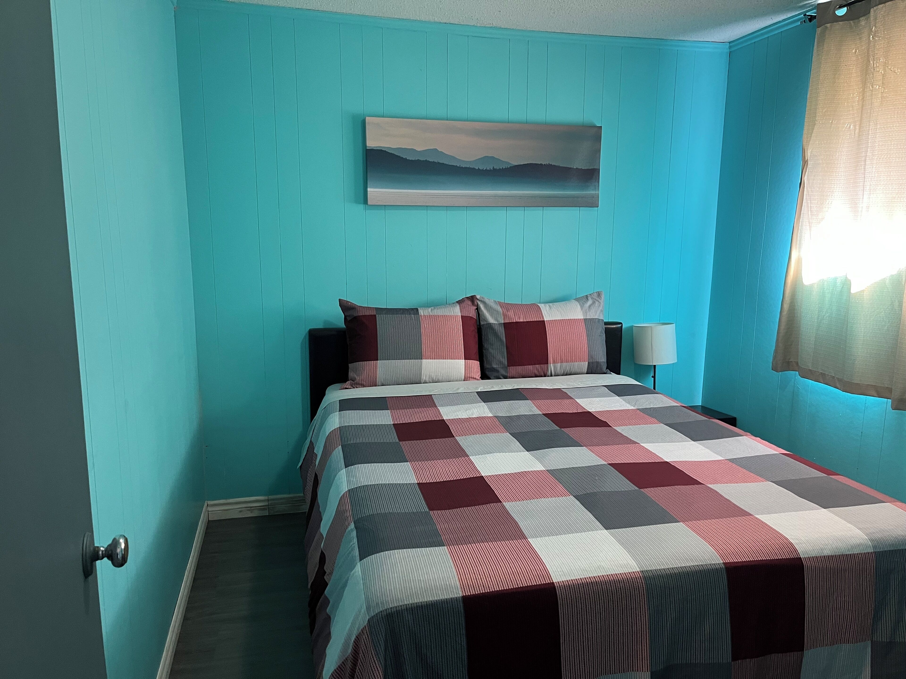 Un dormitorio con paredes color turquesa, una cama con colcha a cuadros, dos almohadas, una mesita de noche con una lámpara y un cuadro en la pared.