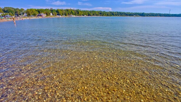 Ein klarer See mit sichtbaren Kieselsteinen und einer entfernten Uferlinie mit Gebäuden und Bäumen.