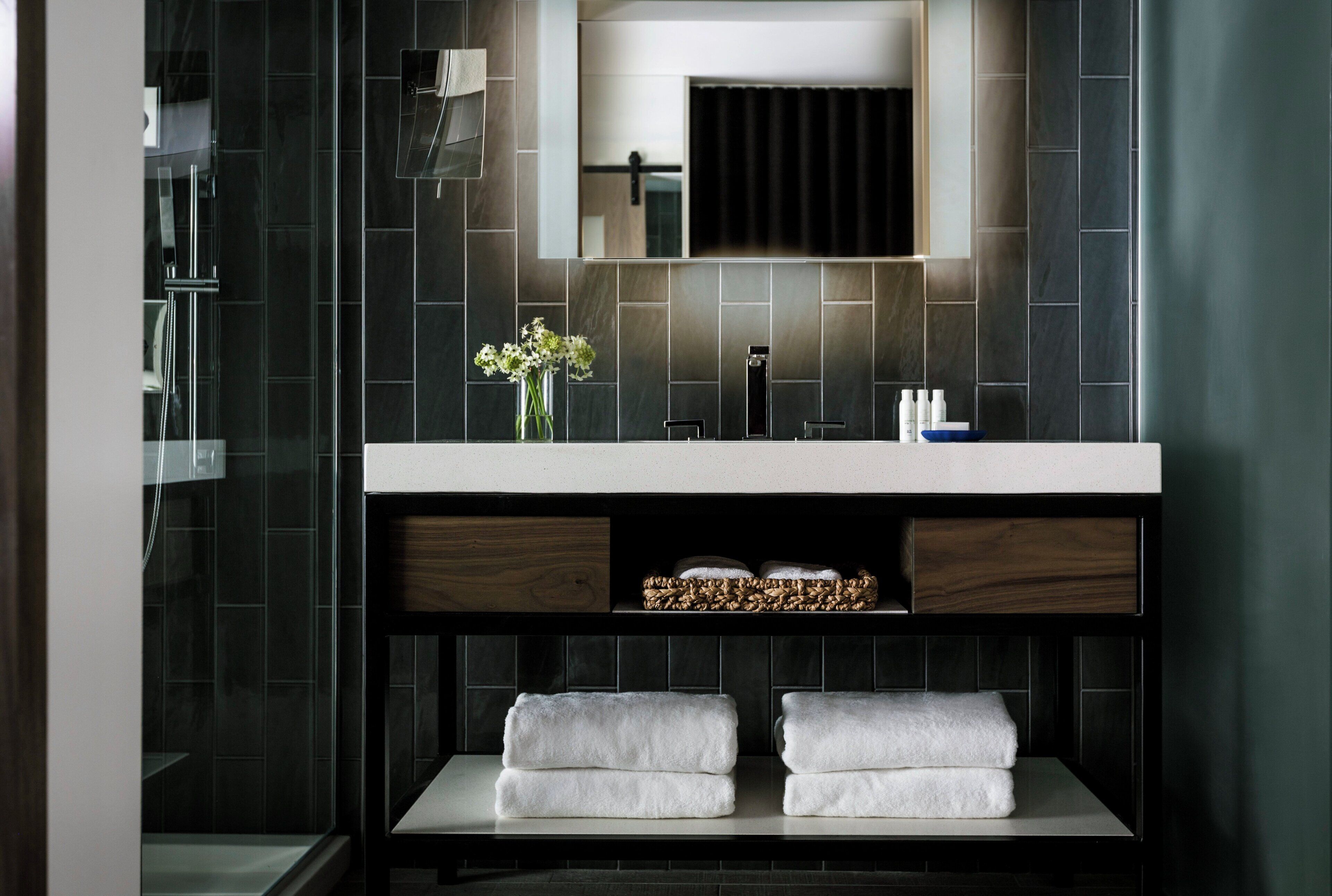 A modern bathroom with a dark tiled wall, a large mirror, a sink, and a shelf with towels.