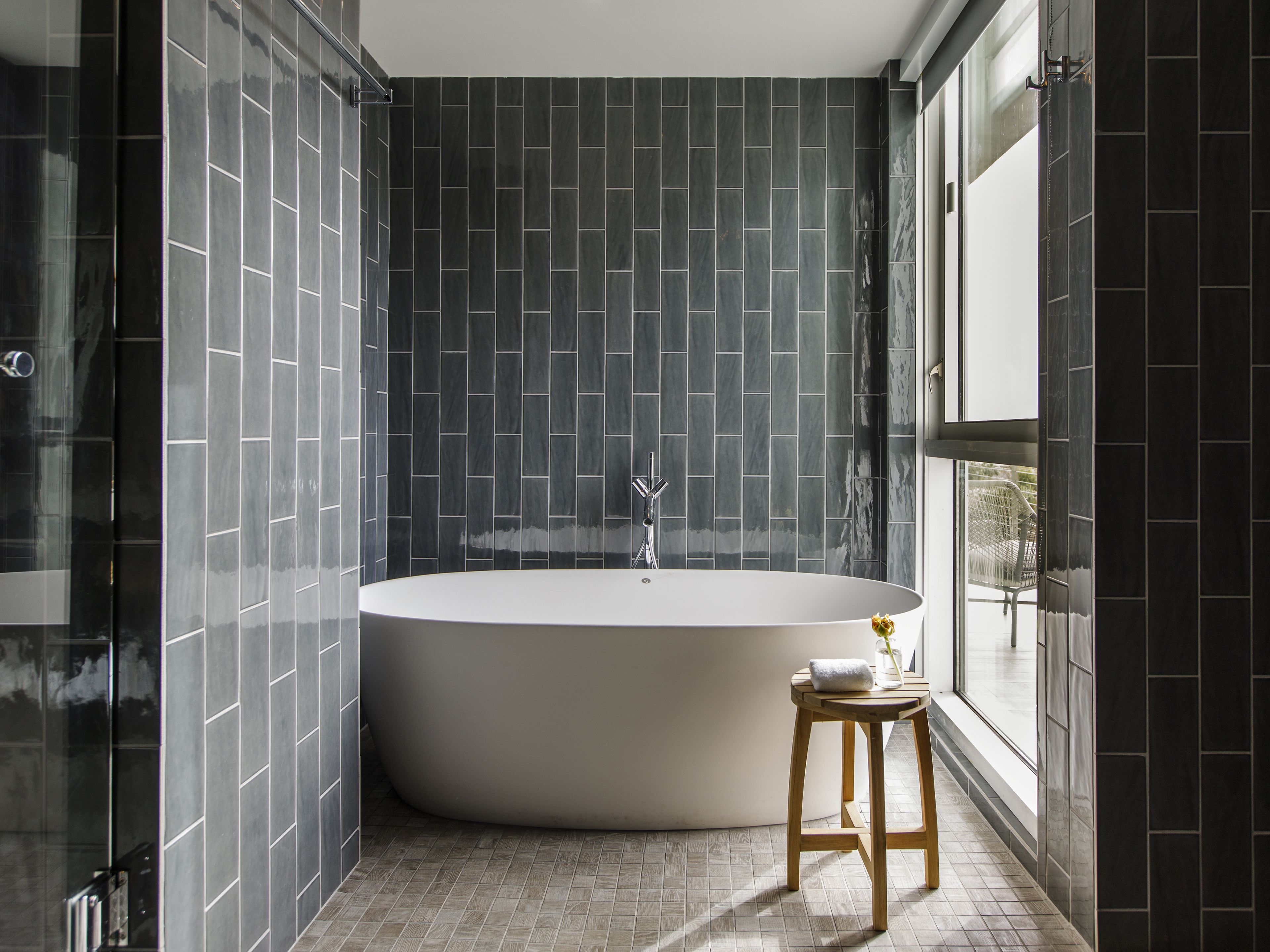 Une salle de bain moderne avec une baignoire sur pieds, des murs recouverts de carreaux foncés et un tabouret en bois.