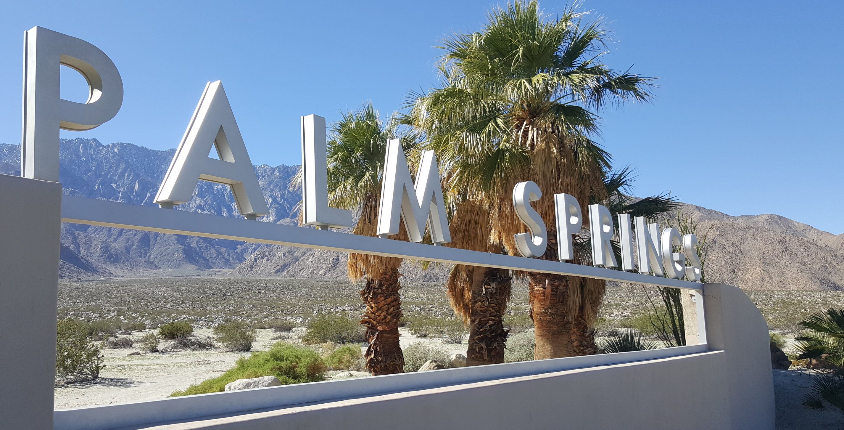 A sign with the text "PALMS SPRINGS" in large letters, set against a backdrop of mountains and palm trees.