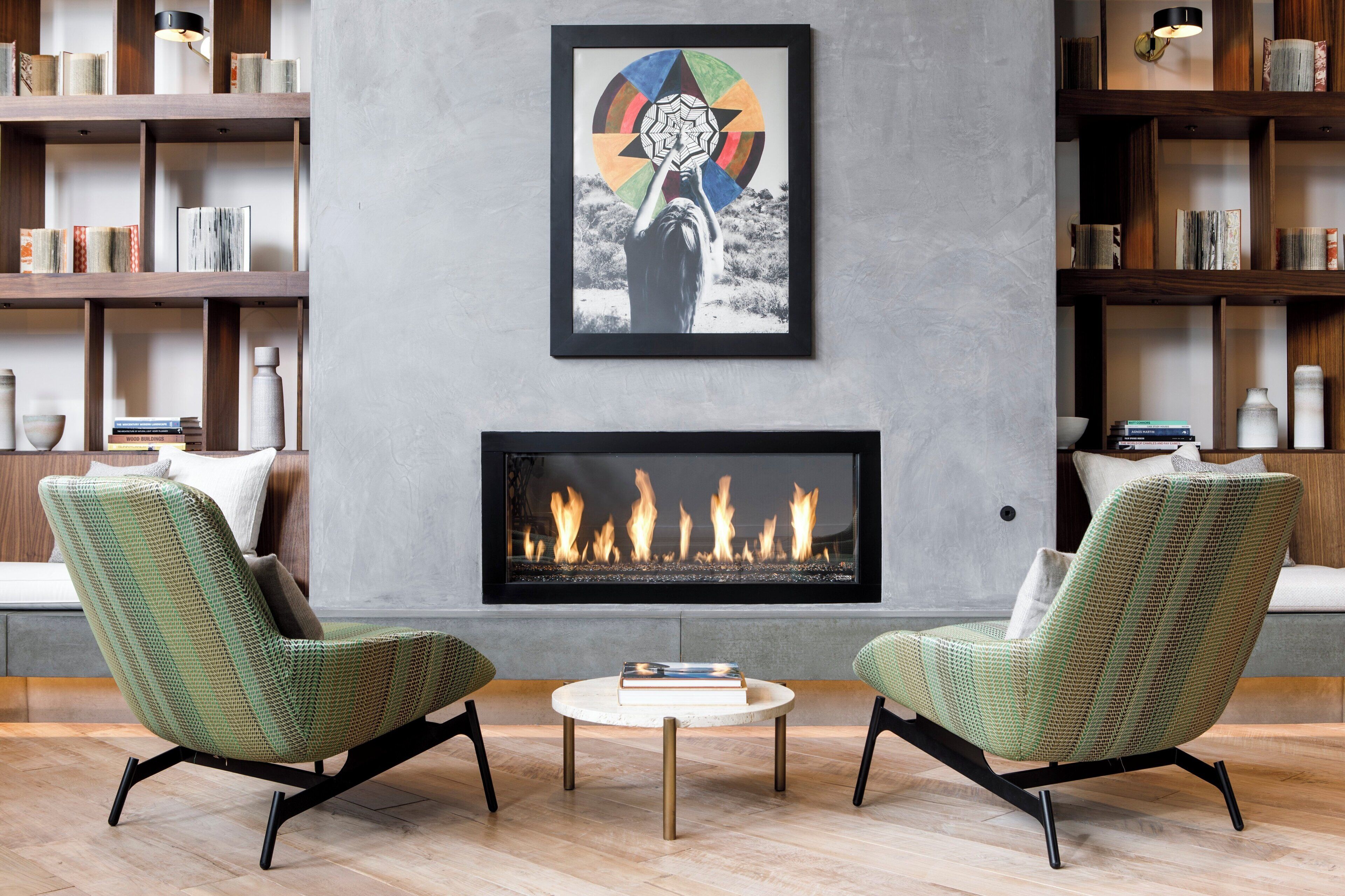 A modern living room with a fireplace, two green armchairs, a coffee table, and bookshelves filled with books.