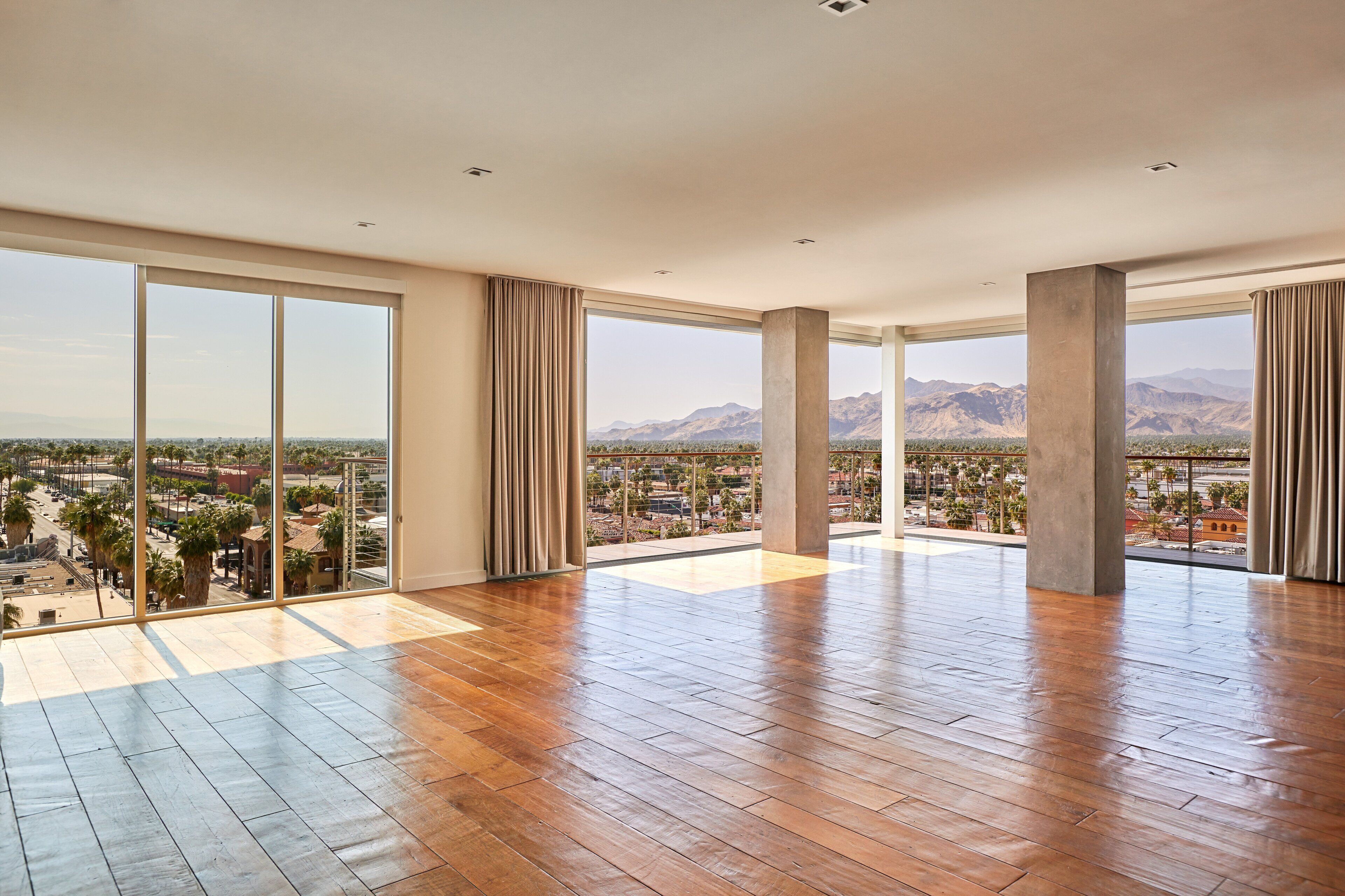 A spacious room with large windows offering a cityscape view, wooden flooring, and a high ceiling.