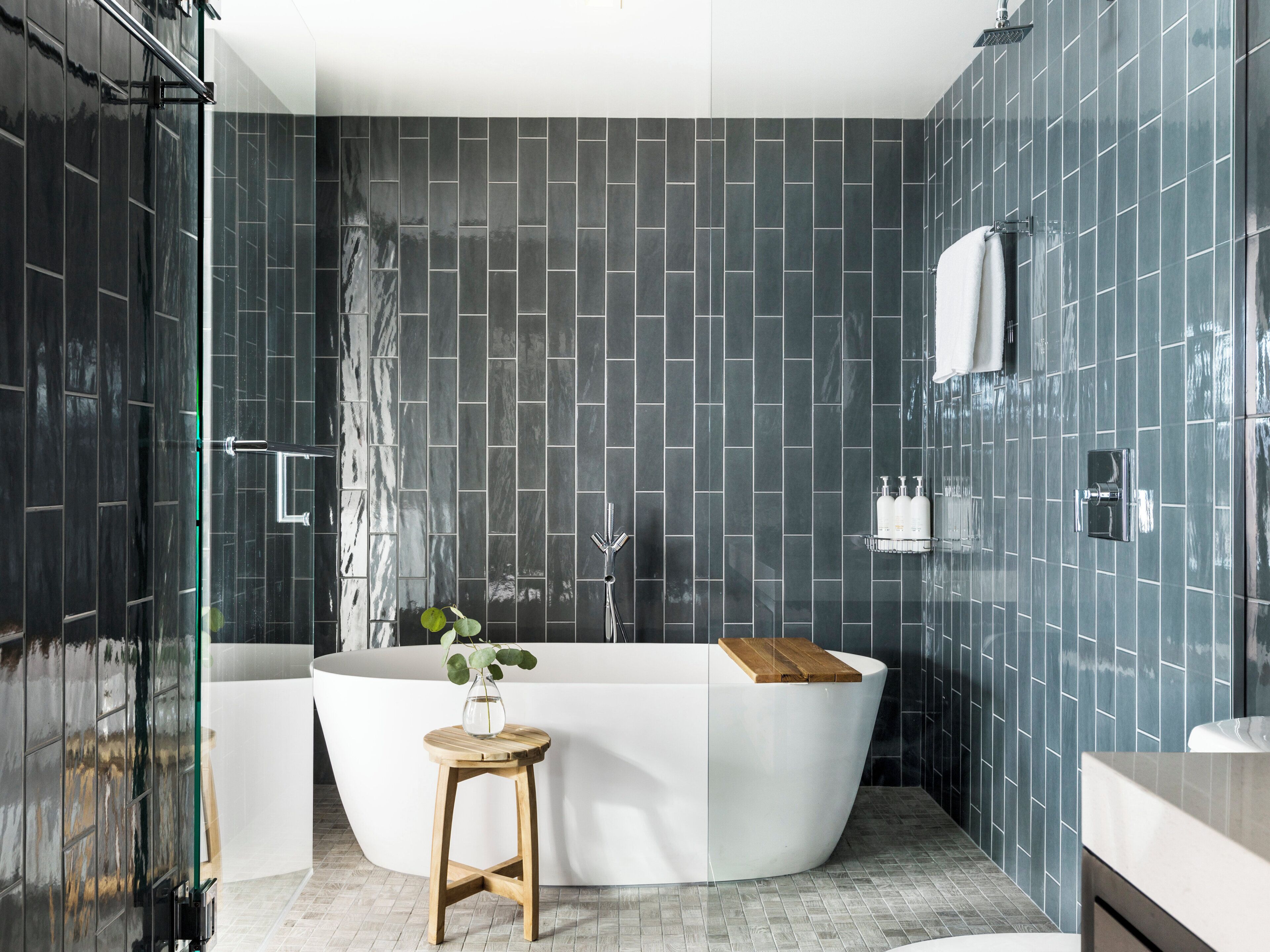 A modern bathroom with dark tiled walls, a freestanding bathtub, a glass shower enclosure, and a wooden stool with a vase.