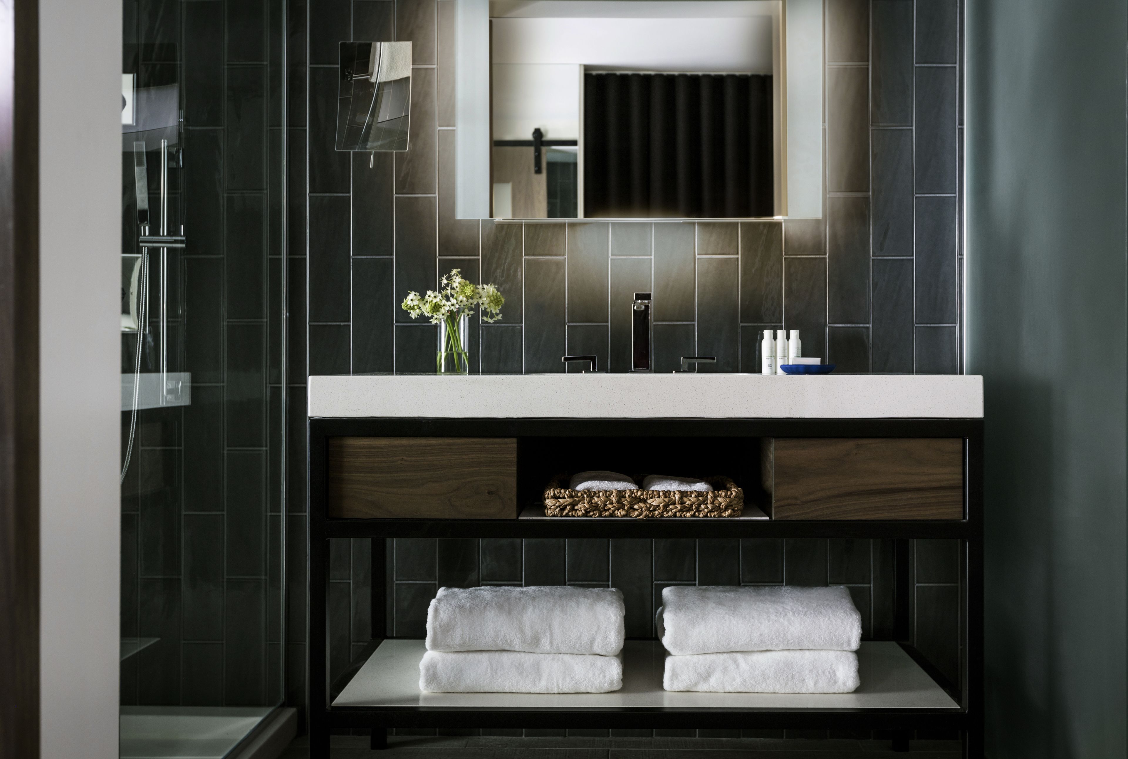 Une salle de bain moderne avec un mur recouvert de carreaux foncés, un grand miroir au-dessus du lavabo et deux serviettes posées sur une étagère en dessous.