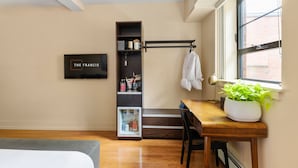 A compact hotel room with a bed, a desk with a chair, a small kitchenette, a bookshelf, and a window with a view of a brick building. - The Francis (Portland)