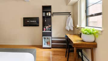 A compact hotel room with a bed, a desk with a chair, a small kitchenette, a bookshelf, and a window with a view of a brick building.