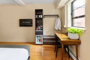 A compact hotel room with a bed, a desk with a chair, a small kitchenette, a bookshelf, and a window with a view of a brick building. - The Francis (Portland)
