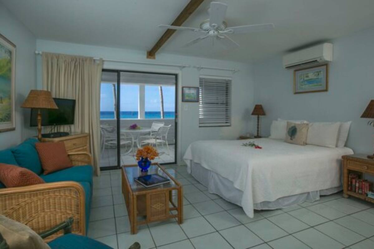 A room with a bed, a sofa, a table with a vase, a ceiling fan, and a view of the ocean.