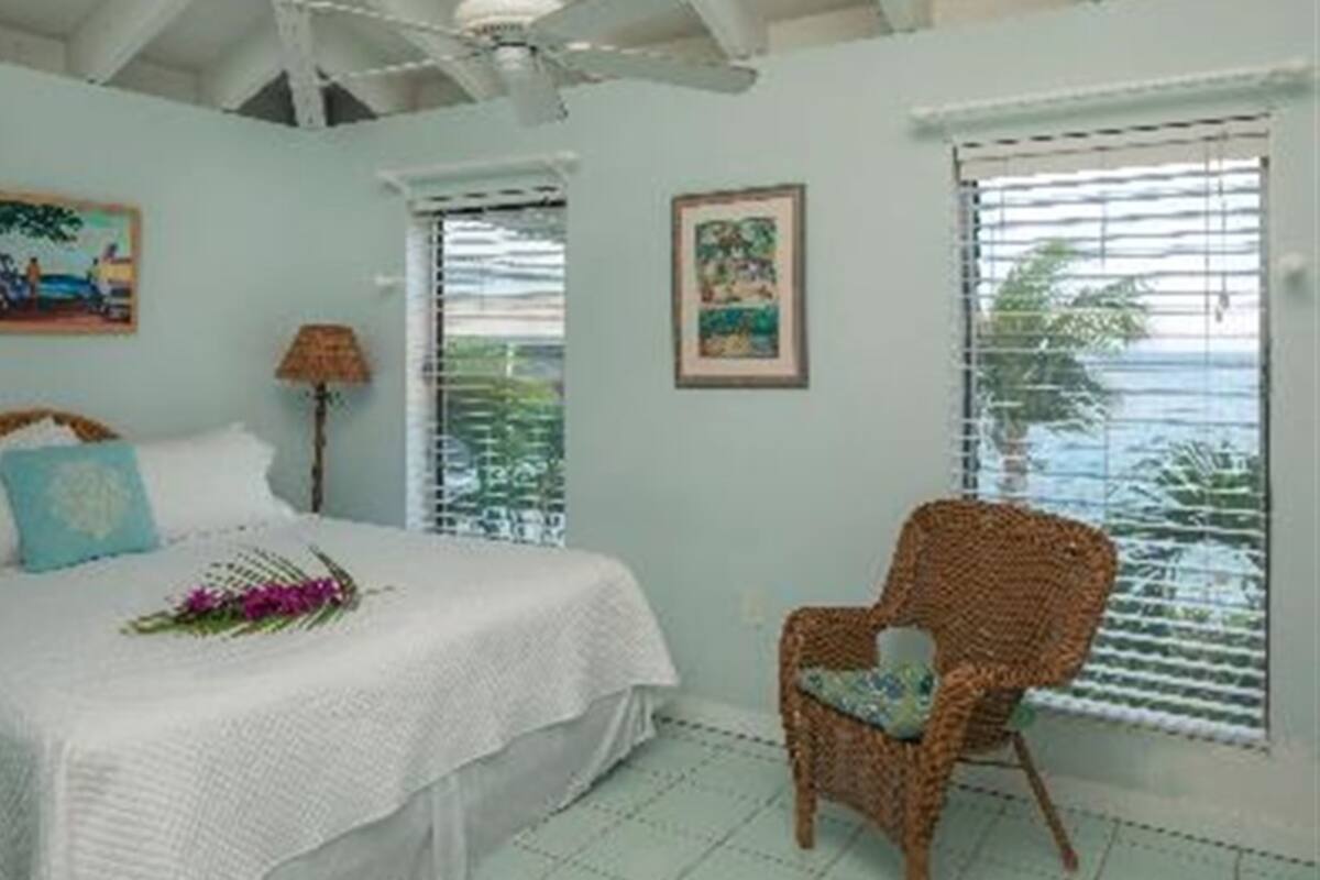 A bedroom with a bed, a wicker chair, and two windows with blinds.