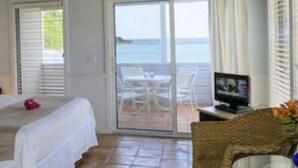 A room with a bed, a TV, a wicker chair, and a balcony with a view of the sea. - Coconut Coast Villas (St. John)