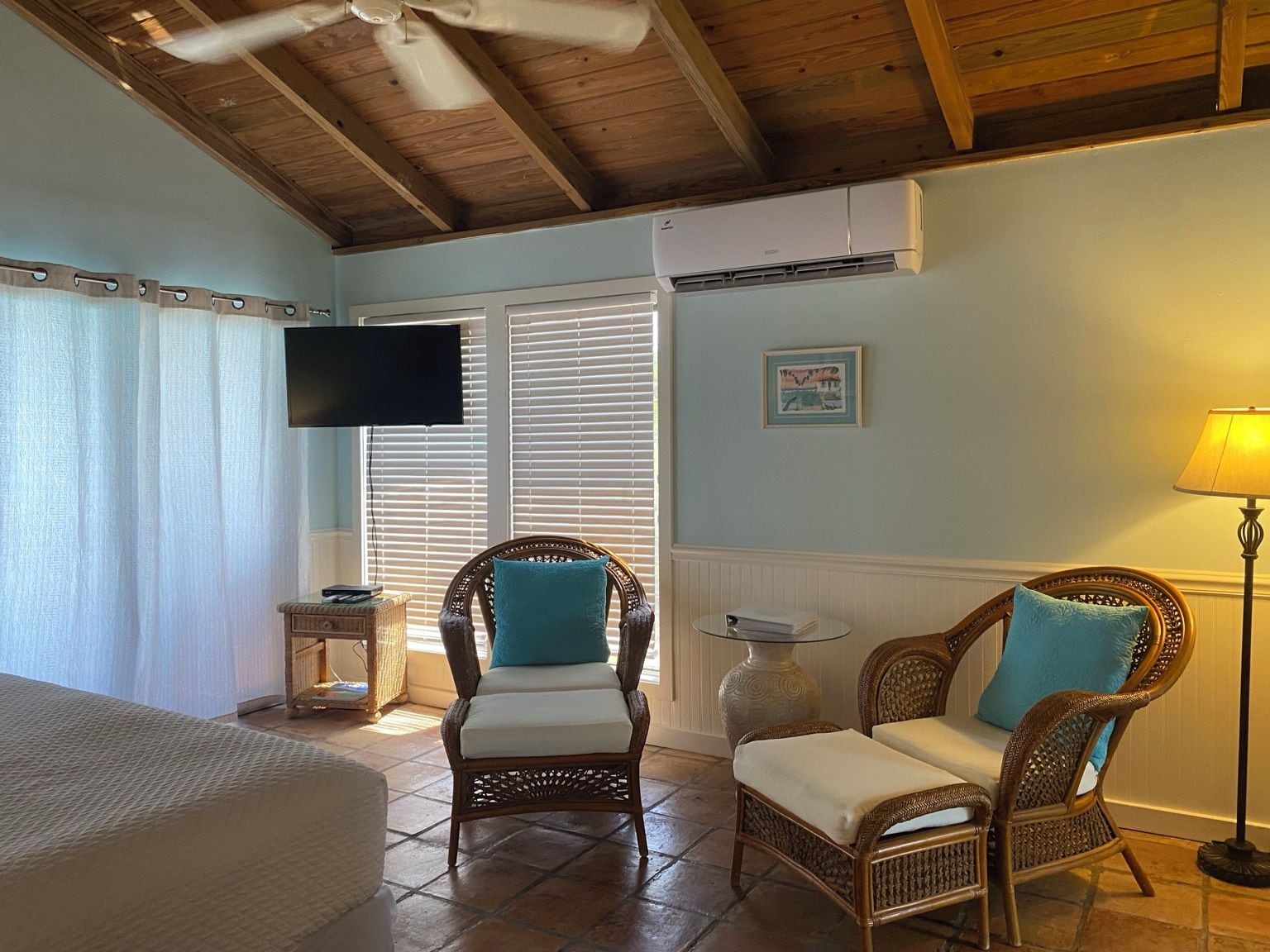 Una stanza con ventilatore a soffitto, televisione a schermo piatto, poltrone in vimini con cuscini, un comodino e una lampada.