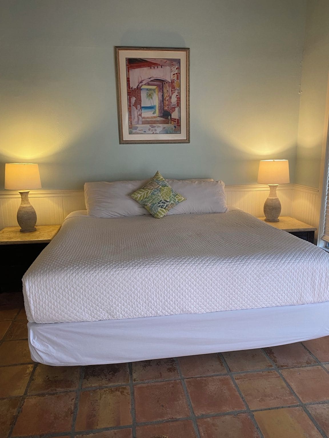 Una cama bien hecha con una colcha acolchada, dos lámparas de noche, una sola almohada y un cuadro en la pared.