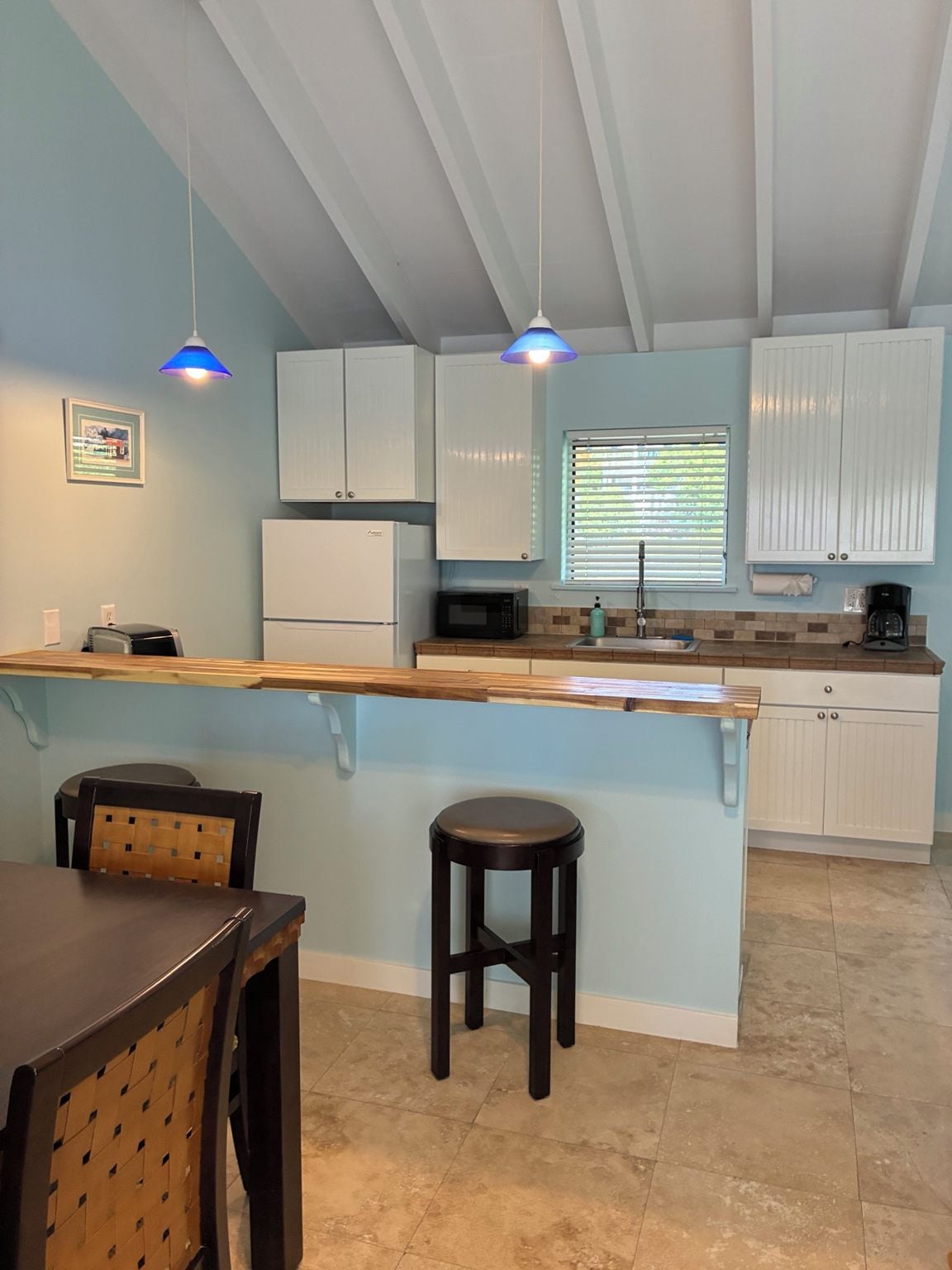 A kitchen with white cabinets, a wooden countertop, a sink, a microwave, and a coffee maker. There are two hanging pendant lights and a window with blinds.
