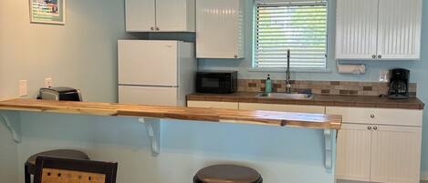 A kitchen with white cabinets, a wooden countertop, a sink, a microwave, and a coffee maker. There are two hanging pendant lights and a window with blinds.