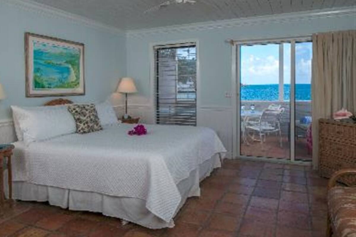 A bedroom with a bed, a nightstand, a lamp, a painting, and a view of the ocean through a sliding glass door.