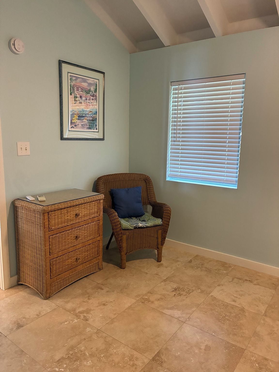 A room with a wicker chest of drawers, a wicker chair with a cushion, a framed picture on the wall, and a window with blinds.