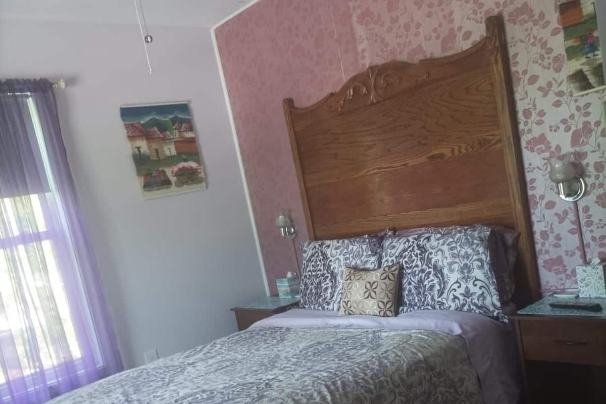 Een slaapkamer met een bed, een houten hoofdbord, een nachtkastje, een plafondventilator, een raam met gordijnen en een bloemen behang.