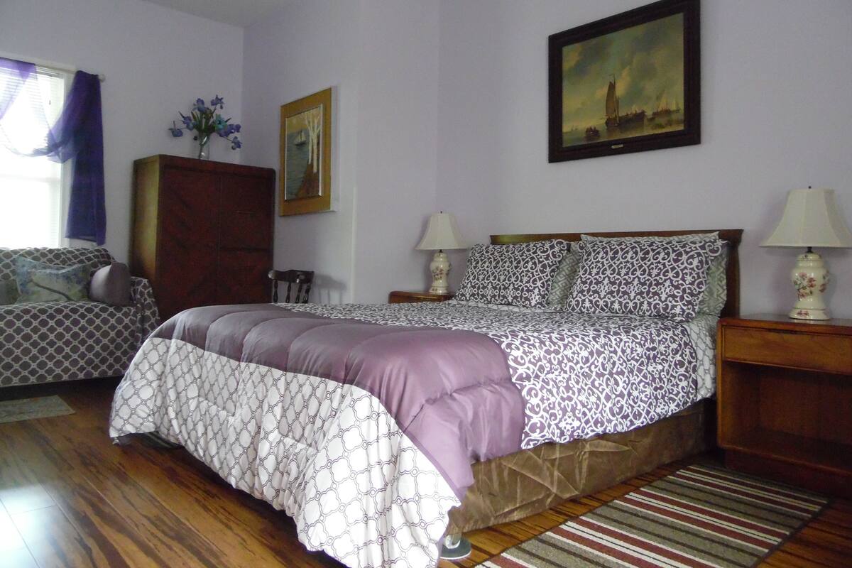 Een slaapkamer met een bed, twee nachtkastjes, een dressoir, een raam met gordijnen, een schilderij aan de muur en een vloerkleed.