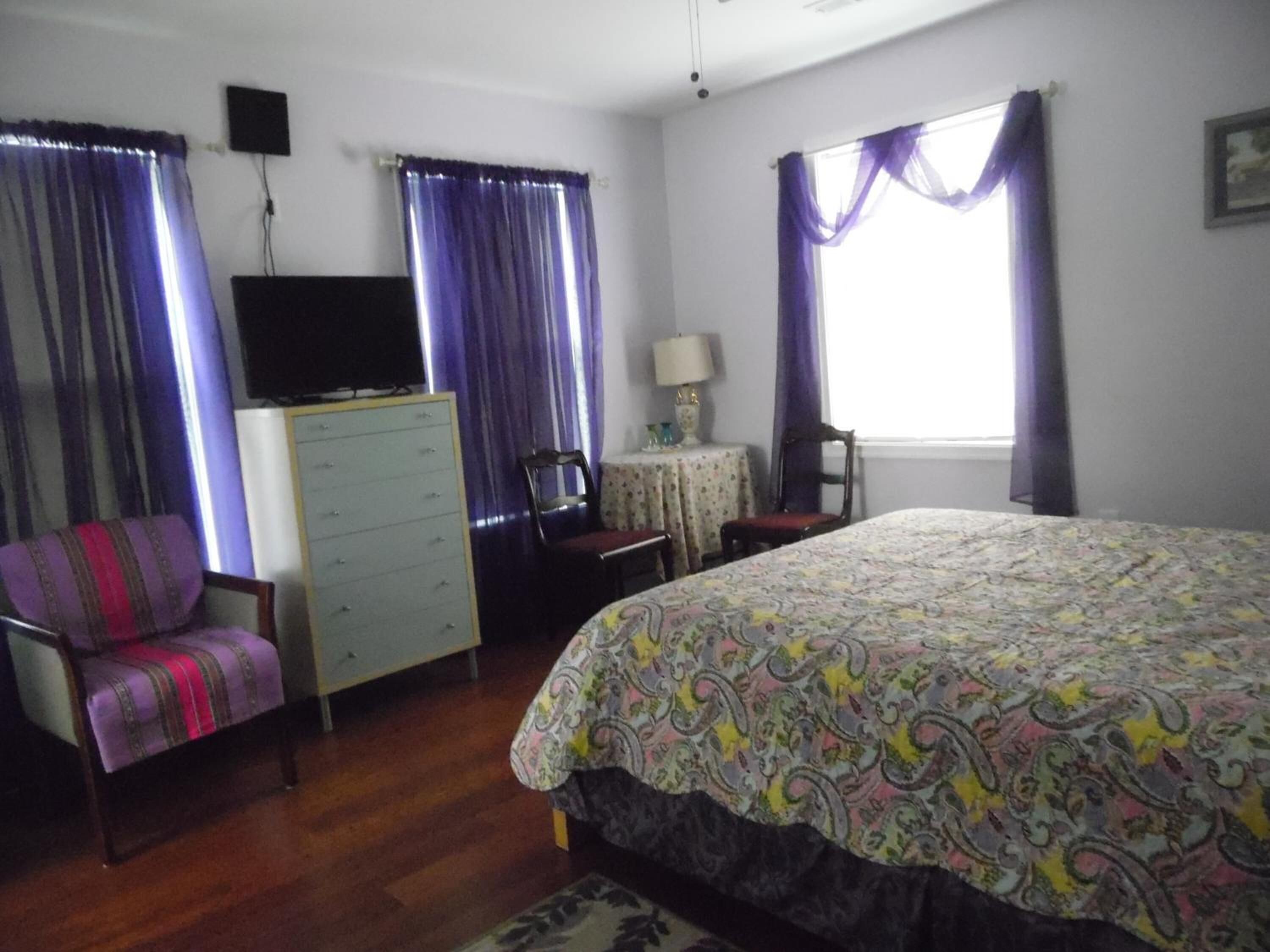 A bedroom with a bed, a dresser, a chair, a TV, and a window with curtains.