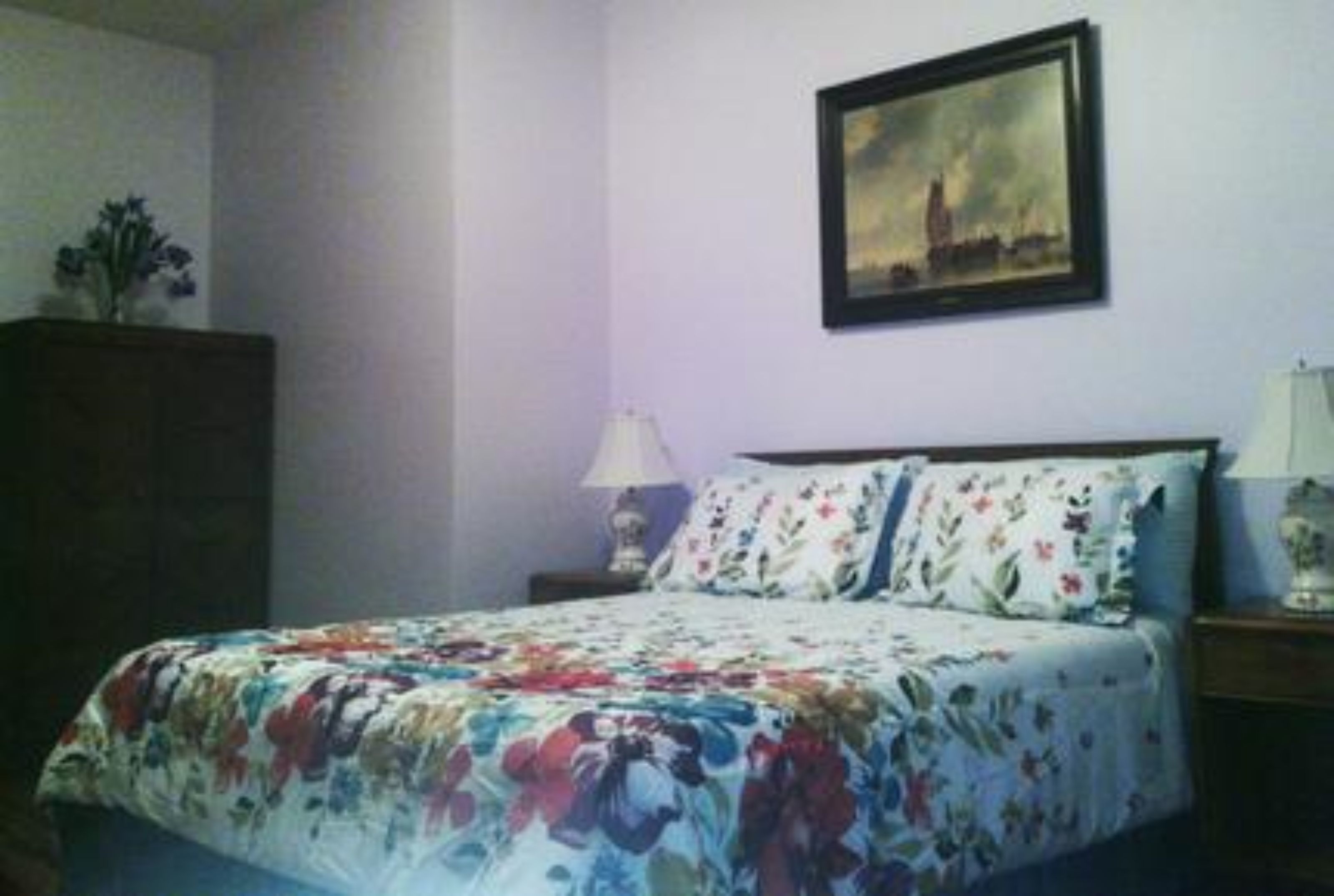 Ein Schlafzimmer mit Blumenmuster-Bettdecke, einem Nachttisch mit Lampe und einem gerahmten Bild an der Wand.