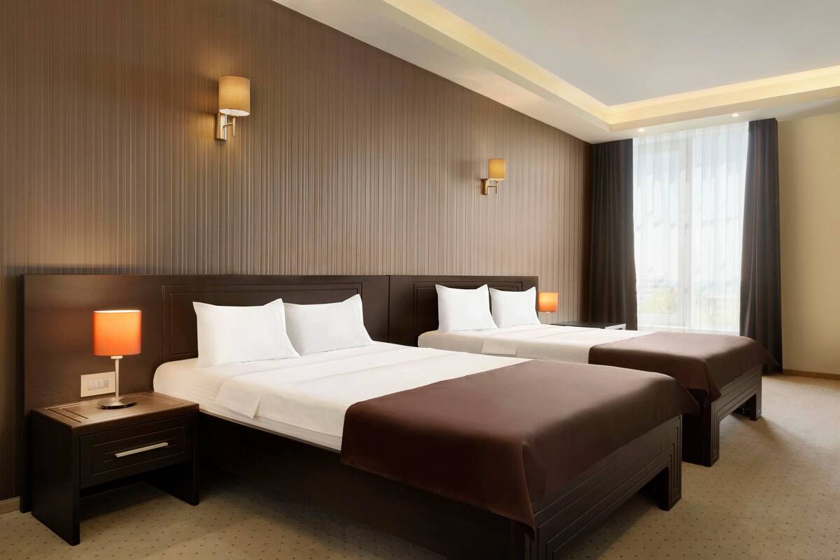 A hotel room with two beds, a nightstand with a lamp, and a large window with curtains.