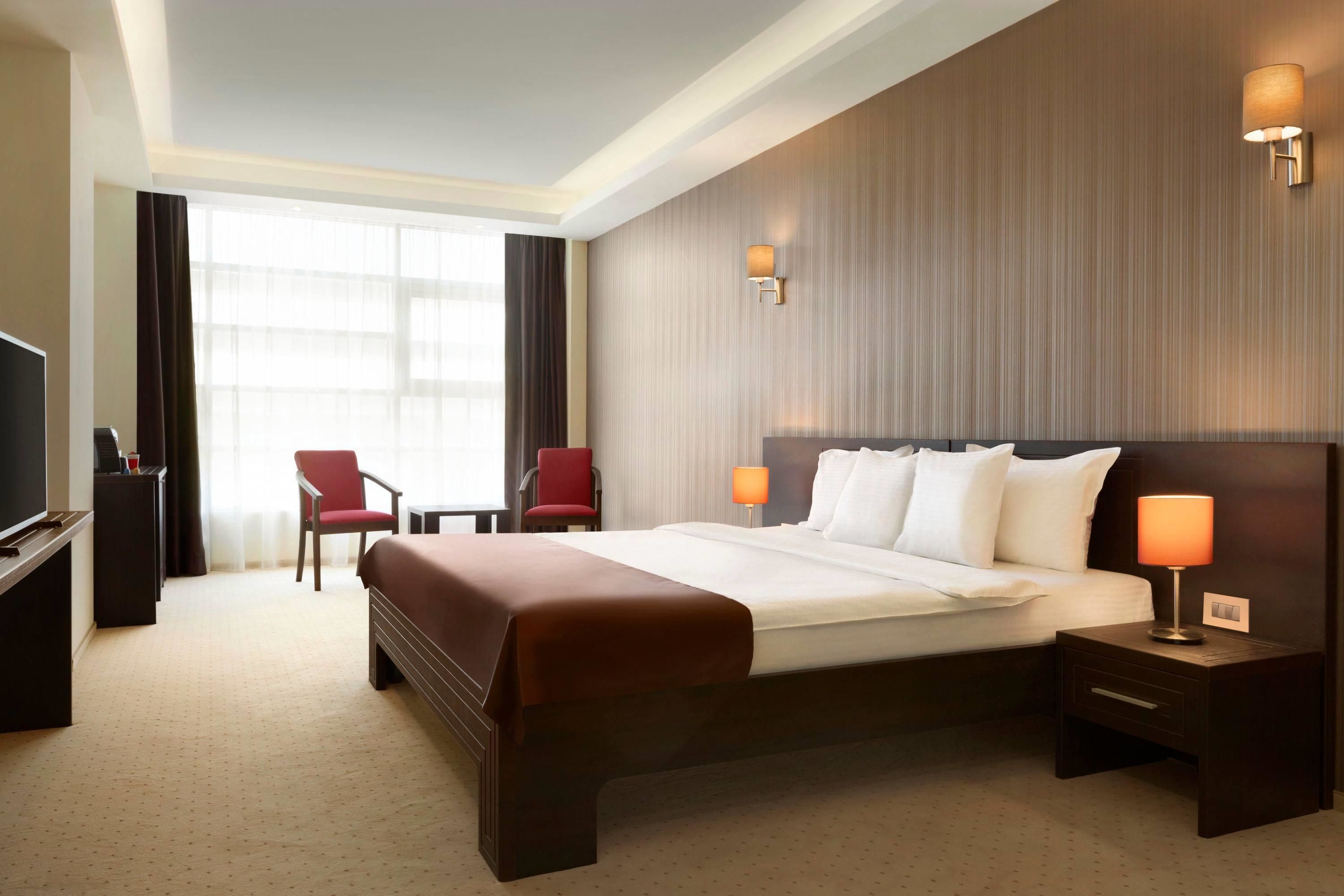 Une chambre d’hôtel avec un grand lit, deux tables de chevet avec des lampes, une télévision, un fauteuil rouge et un bureau avec une lampe.