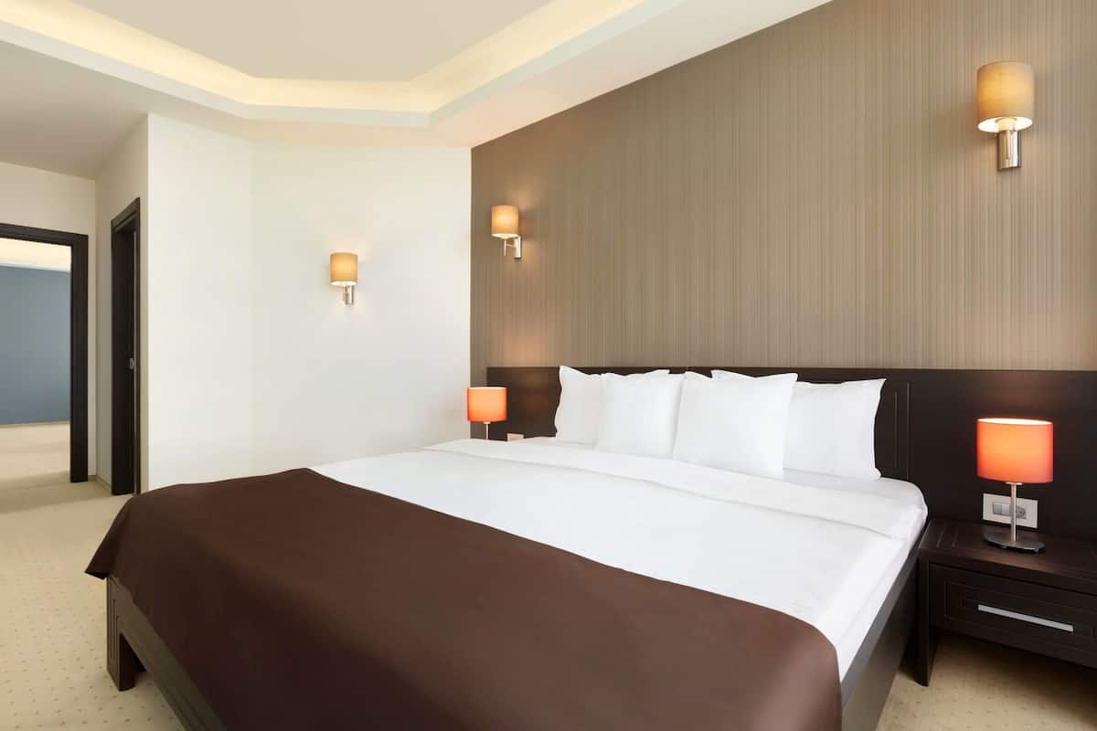 A hotel room with a large bed, two bedside lamps, a wooden headboard, and a view of the sea through an open door.