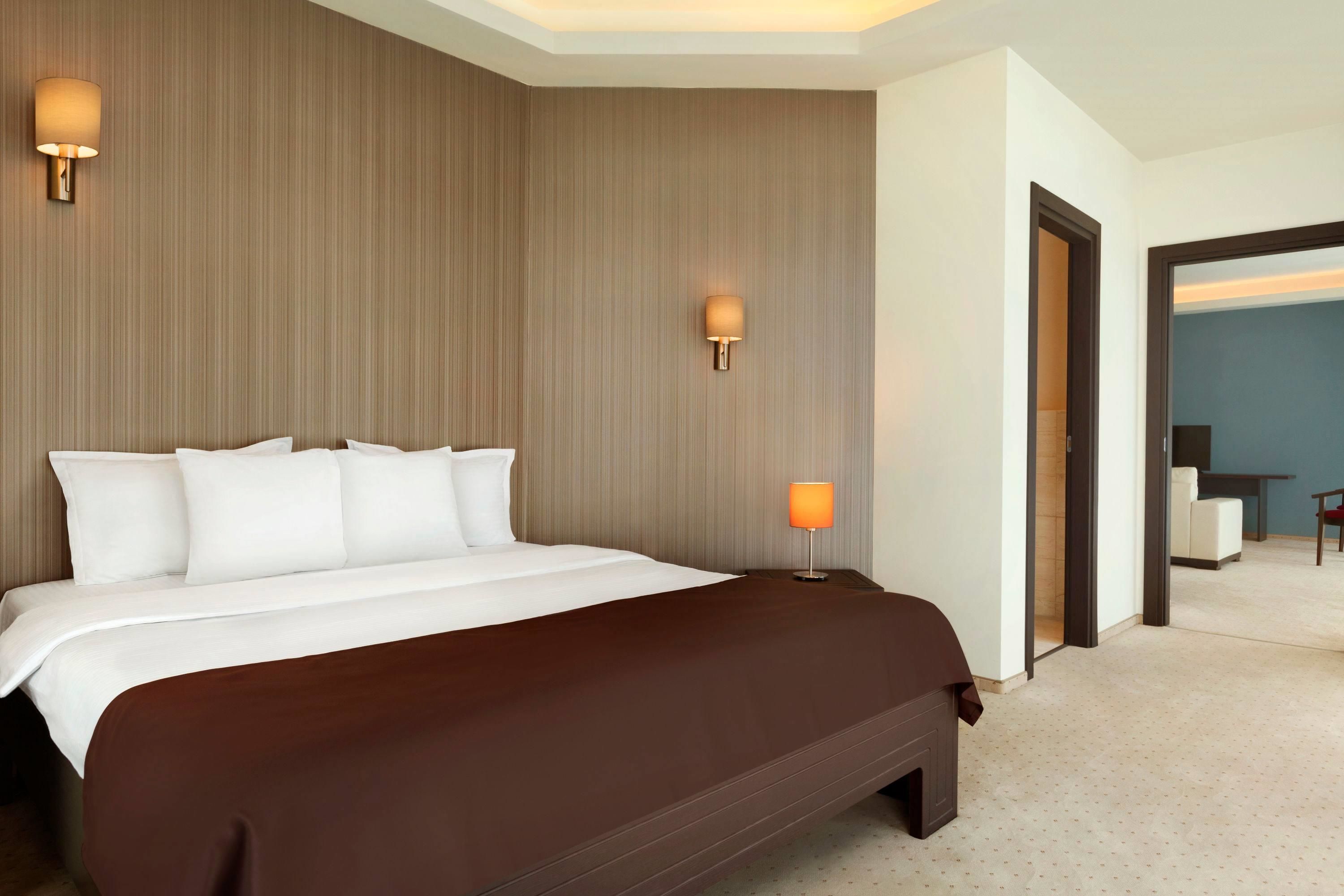 A hotel room with a large bed, two bedside lamps, a wooden headboard, and a view of a dining area through an open door.