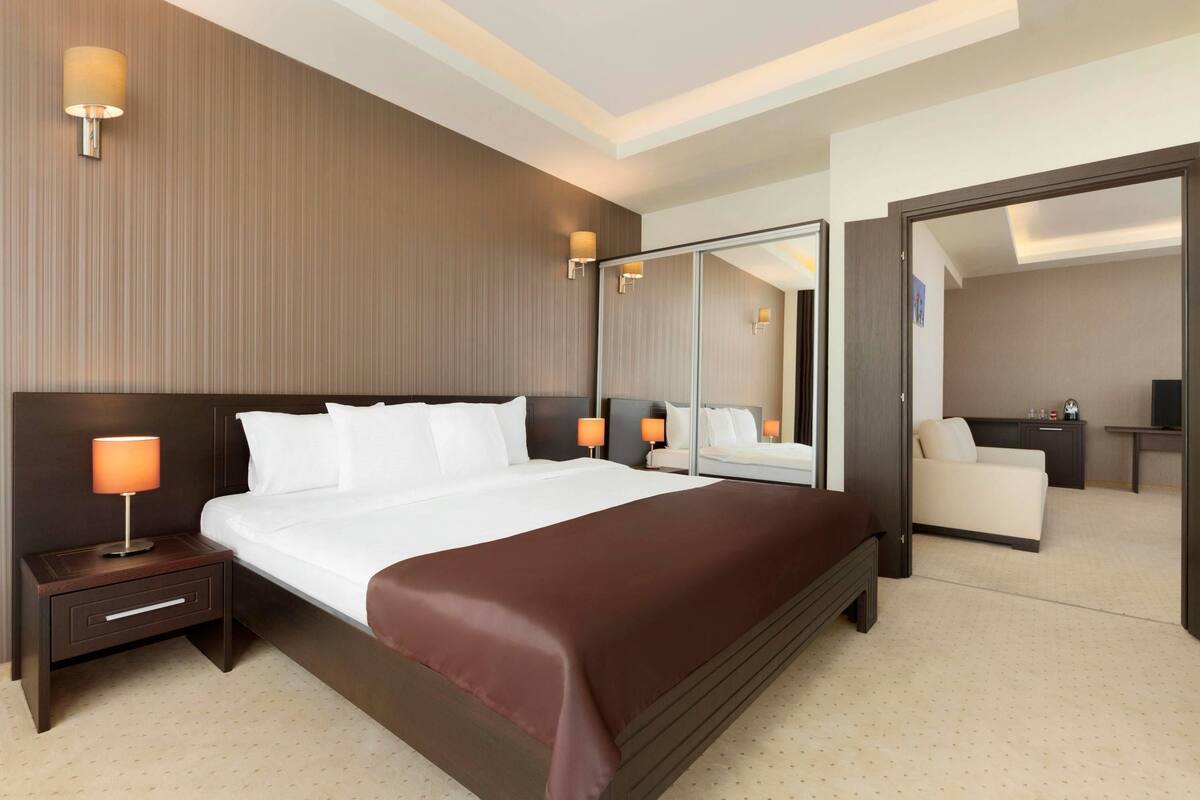 A modern hotel room with a large bed, a nightstand with a lamp, a mirror, and a seating area with a sofa and a table.
