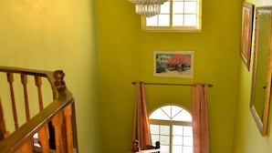 A staircase with wooden railings leading to a room with yellow walls, a large arched window, and a chandelier. - Chateau Margarite Hotel (Montego Bay)