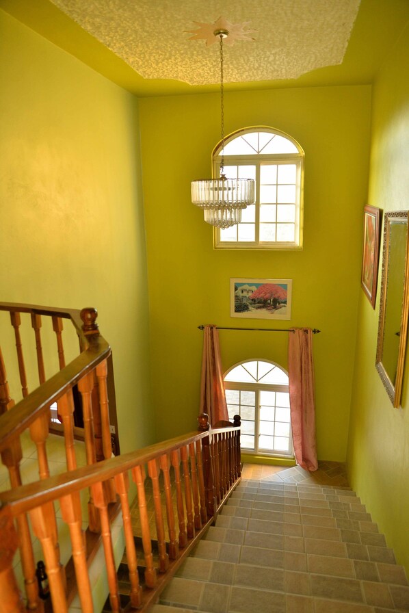A staircase with wooden railings leading to a room with yellow walls, a large arched window, and a chandelier. - Chateau Margarite Hotel (Montego Bay)