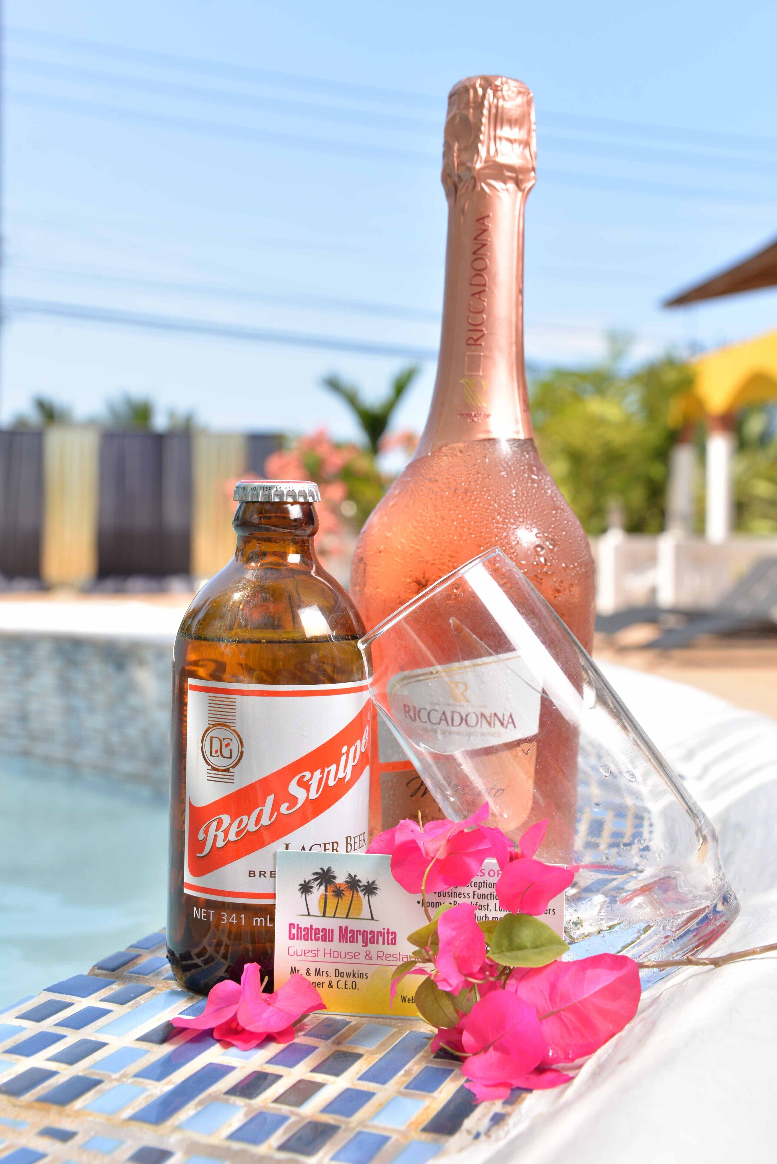 Una botella de cerveza Red Stripe, una botella de vino rosado y un vaso sobre una mesa junto a una piscina.