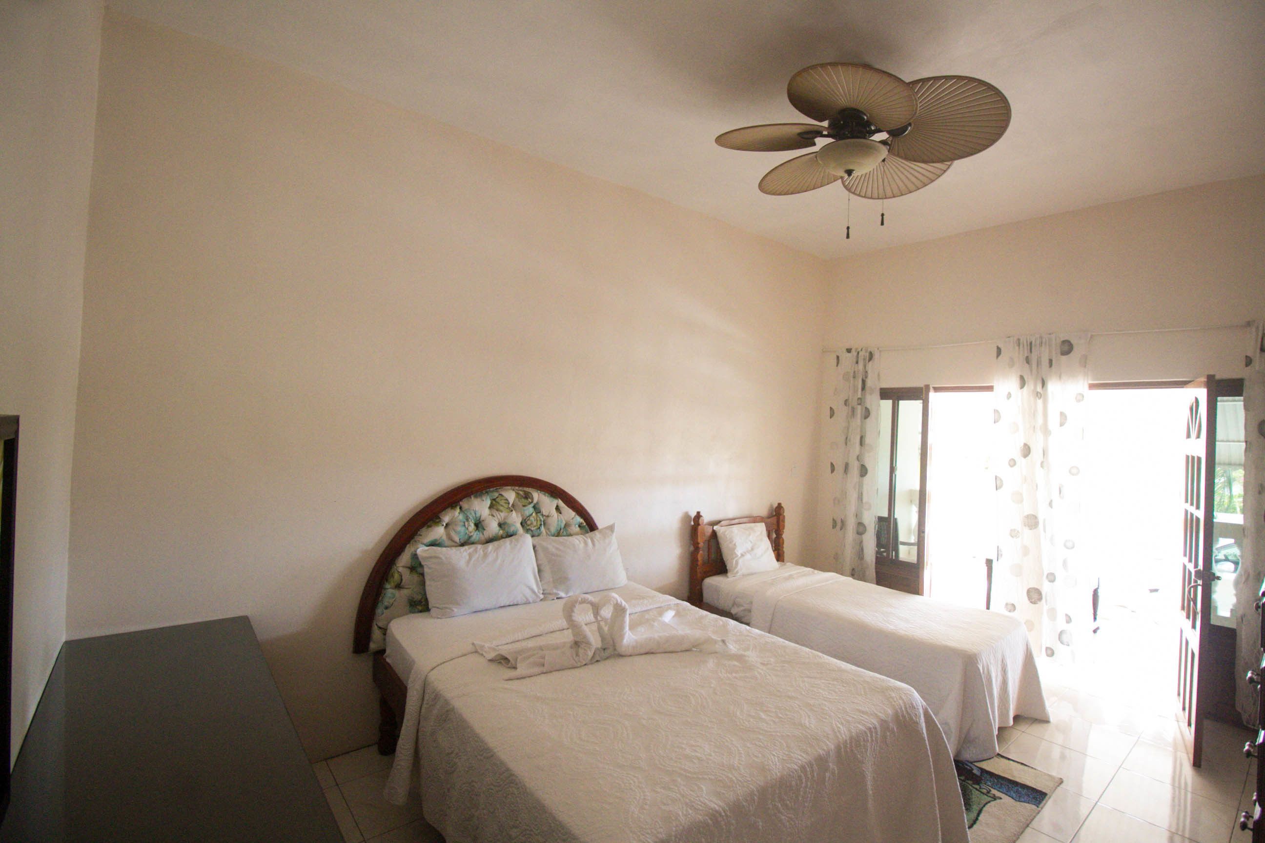 A hotel room with two beds, a ceiling fan, and a view of the outdoors through a door with curtains.