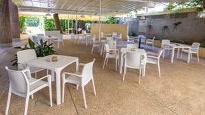 Zona de pati a l'aire lliure amb taules i caires de plàstic blanc, envoltada de vegetació i una paret de pedra.