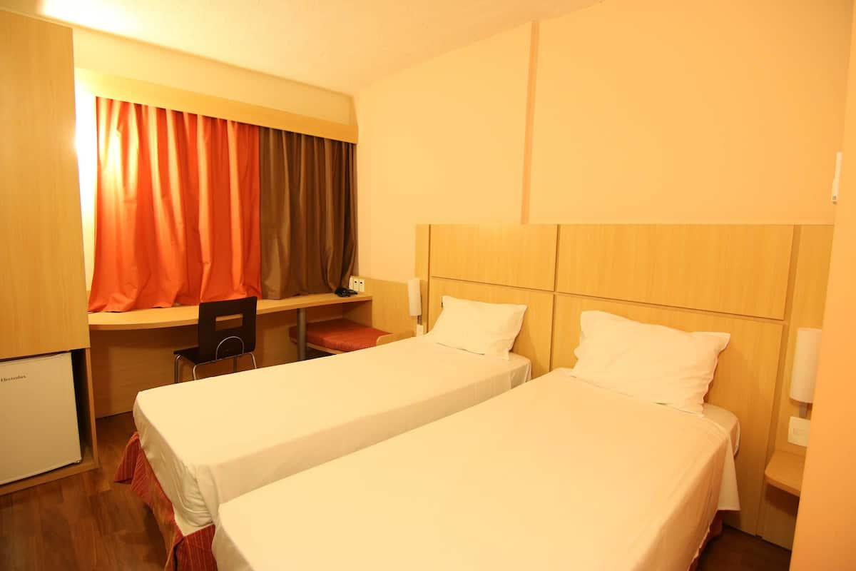 A hotel room with two single beds, a desk, a chair, and a small refrigerator.