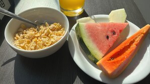 A table with a bowl of cereal, a glass of orange juice, and a plate with watermelon and papaya slices. - Samba Itaboraí (Itaborai)