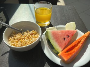 Una mesa con un tazón de cereales, un vaso de jugo de naranja, y un plato con trozos de sandía y papaya.