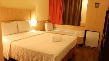 Ein Hotelzimmer mit einem großen Bett, einem Nachttisch, einem kleinen Kühlschrank und einem Fenster mit Vorhängen.