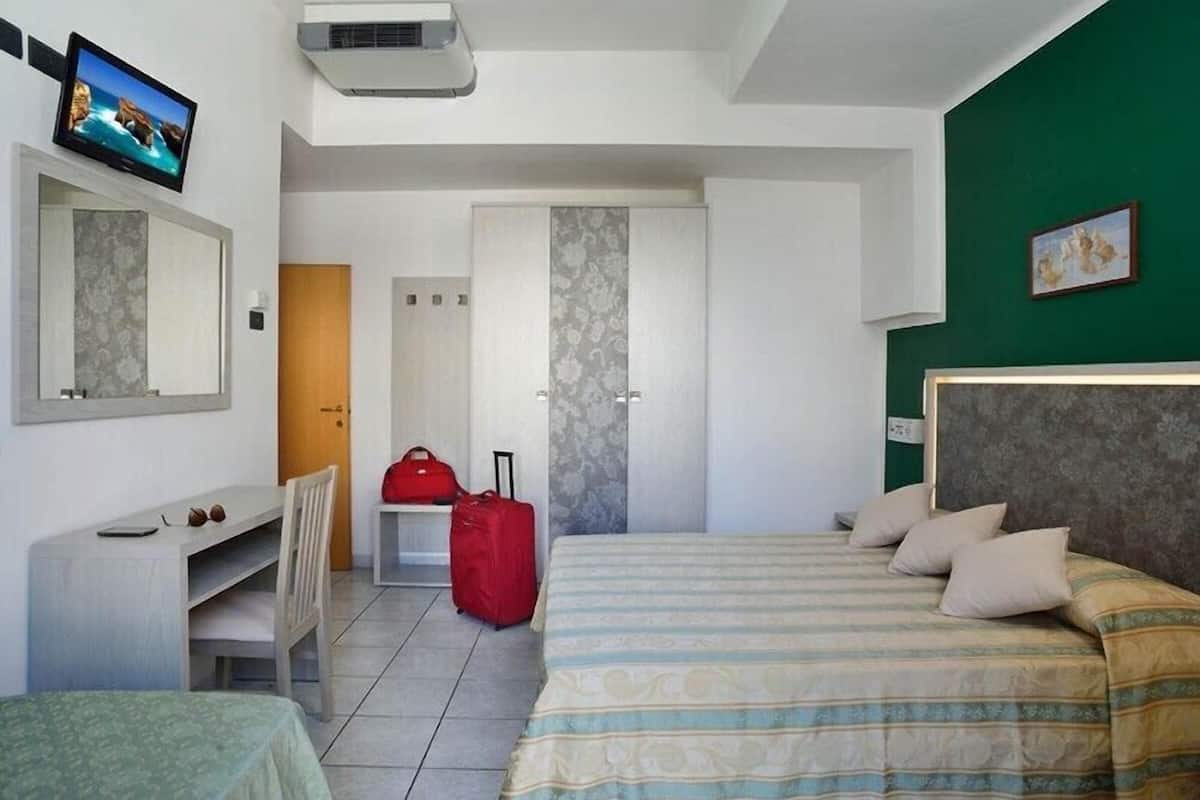 A hotel room with a bed, a desk, a chair, a red suitcase, and a television on the wall.