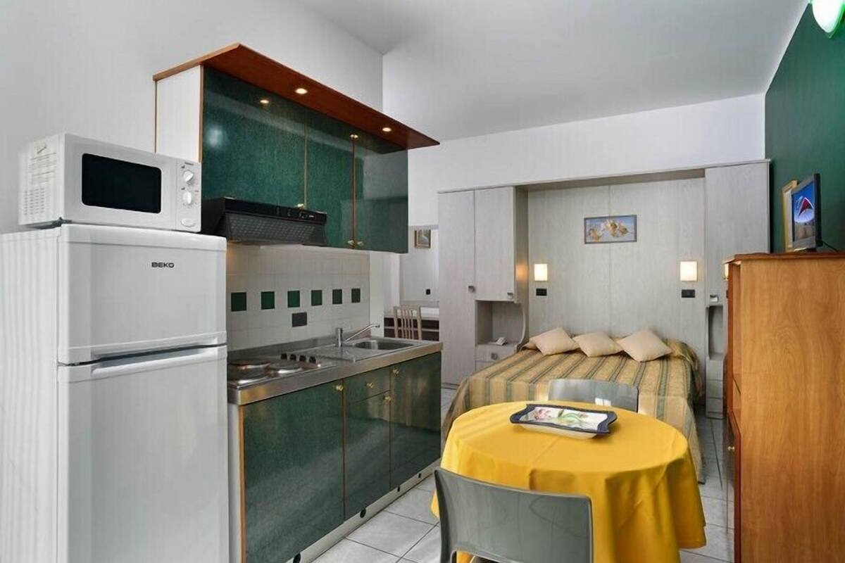 A compact kitchen with a microwave, stove, and sink. There is a dining area with a yellow table and chairs. The room has a bed with a striped bedspread and a small painting on the wall.