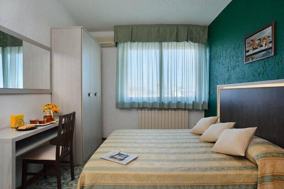 A bedroom with a bed, a desk, a chair, a radiator, and a window with curtains.