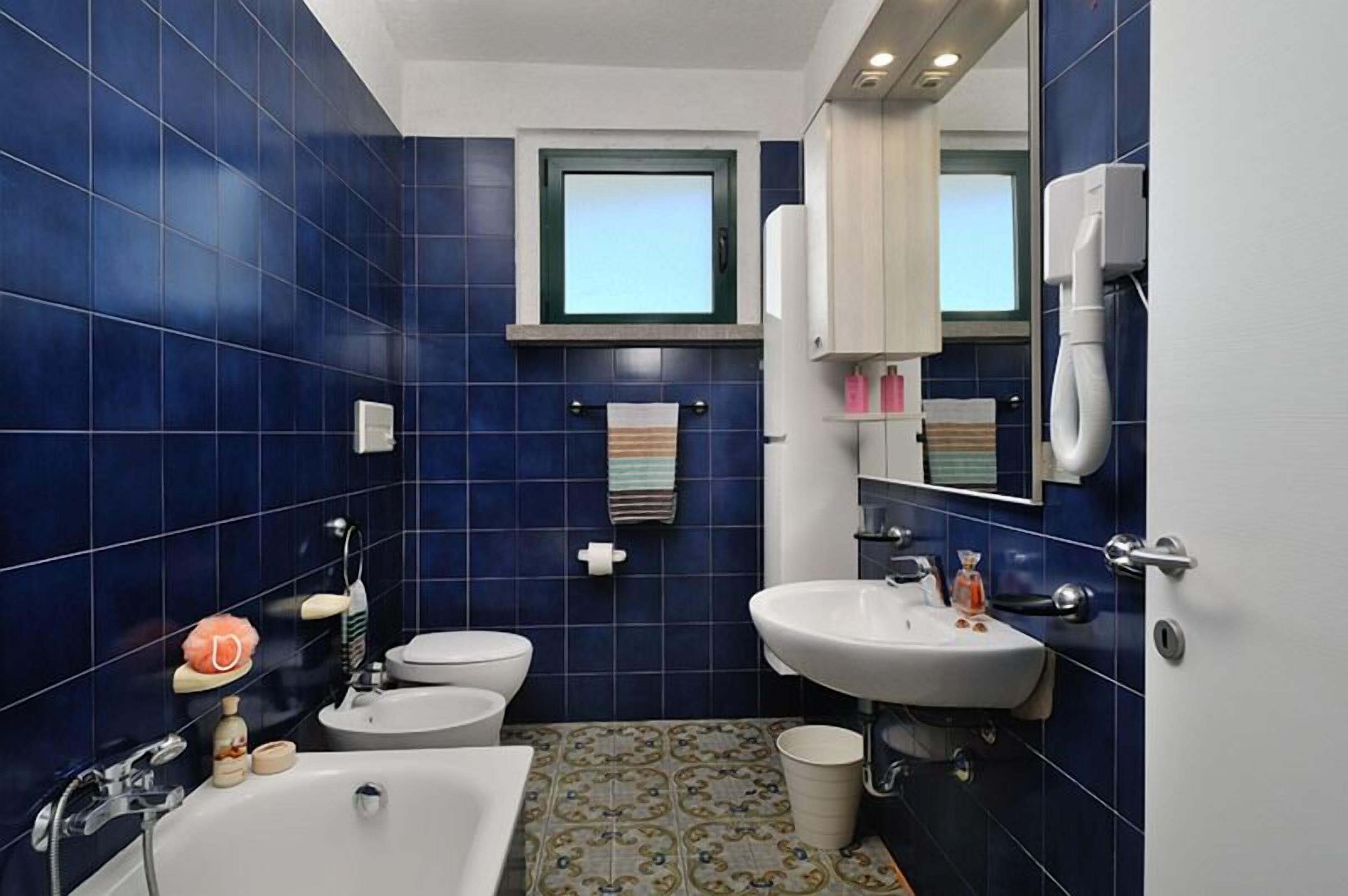 Un bany amb rajoles blaves, un lavabo blanc, un vàter i una banyera.